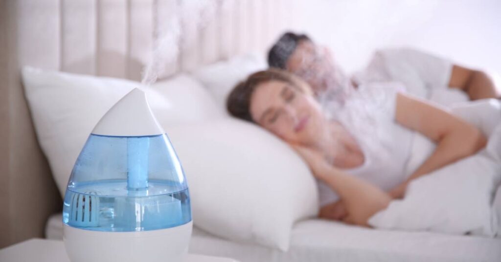 In the foreground, a portable humidifier sits on a nightstand and emits steam. Two people rest in bed in the background.
