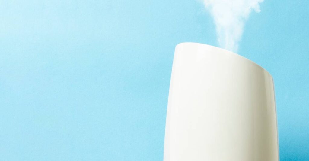 The close-up of a portable white humidifier as it emits steam into the air against a light blue background.