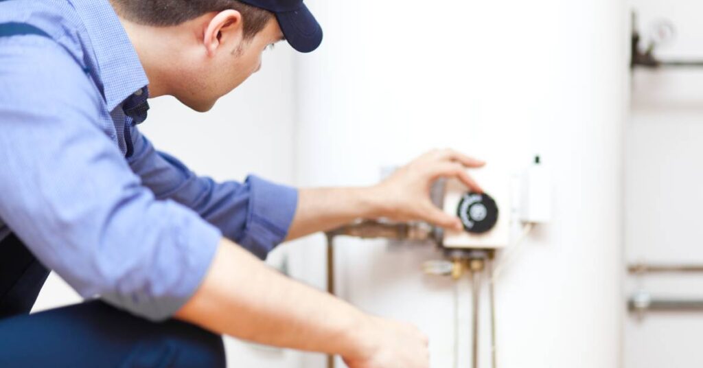 A person in a blue worker's uniform and cap touching the temperature control dial of a water heater with their left hand.