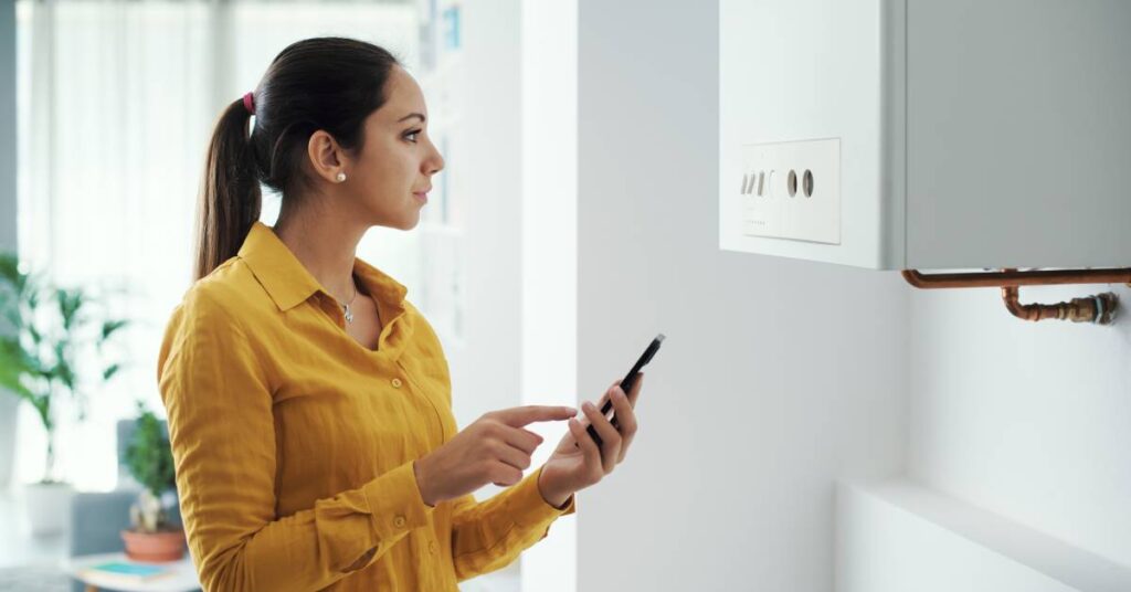 A young woman holding her smartphone in her left hand as she examines what appears to be a smart water heater or boiler.