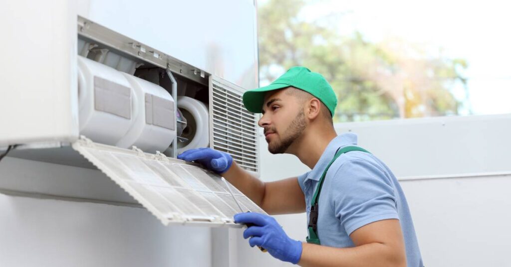 The image of a professional technician in gloves and a cap, looking into the internal parts of an HVAC system.