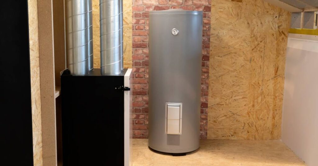A grey electric water heater in front of a brick and wooden wall. Nearby is a box with metal pipes connected to it.