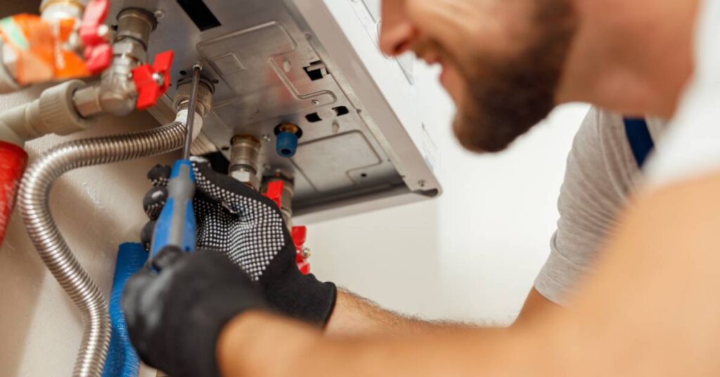 A close-up of a smiling worker installing a water heater. He's wearing black gloves and using a blue screwdriver.