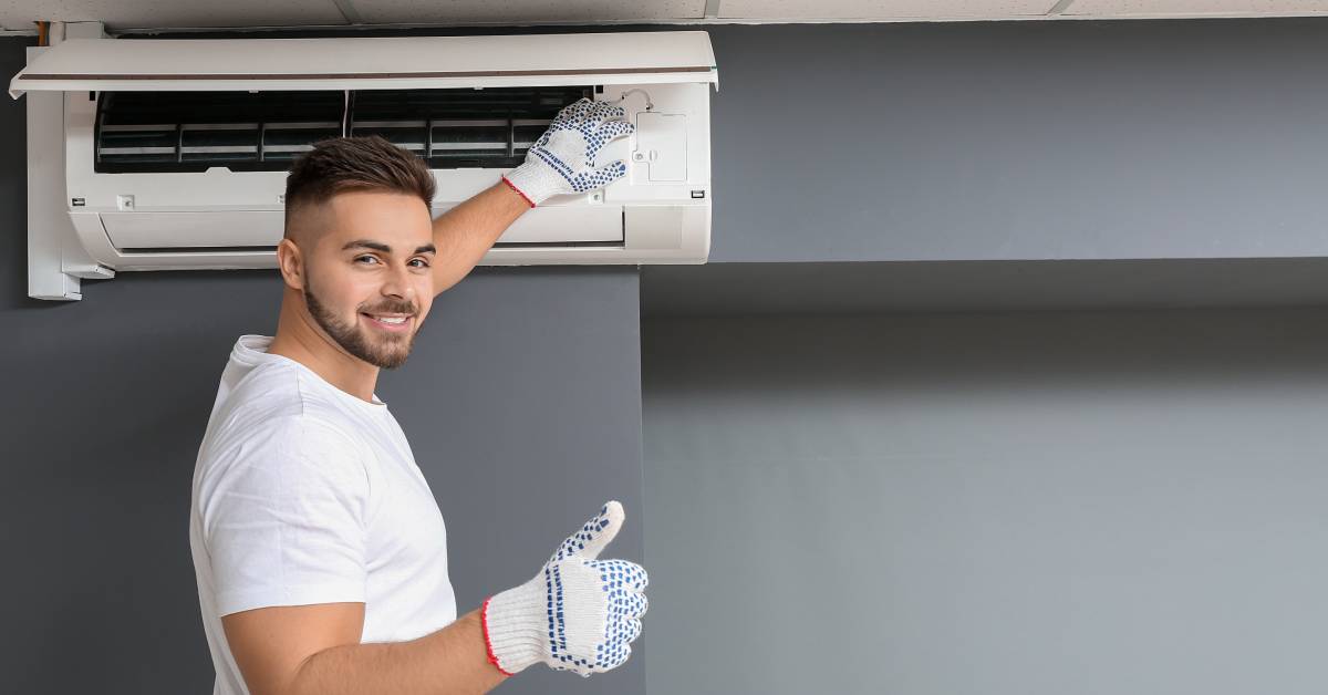 A man smiles, looks toward the camera, and touches an indoor air conditioner. He wears gloves and holds his thumb up.