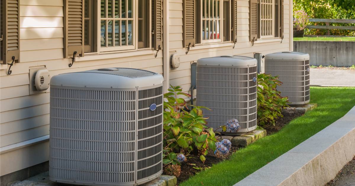 Three air conditioning units near windows at the back of a residential property. The units sit on a stone surface.