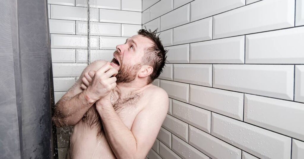 A man stands in the shower and looks at the showerhead with a shocked expression. He holds his arms to his chest.