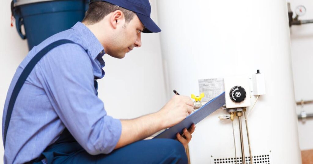 A worker squats near a water heater and writes on a clipboard. He wears a light blue shirt, dark blue pants, and a hat.