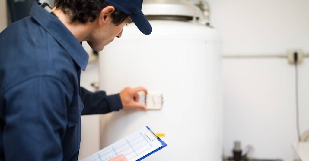 A person wearing a blue uniform and cap holds a clipboard in his right hand and touches a water heater with his left.