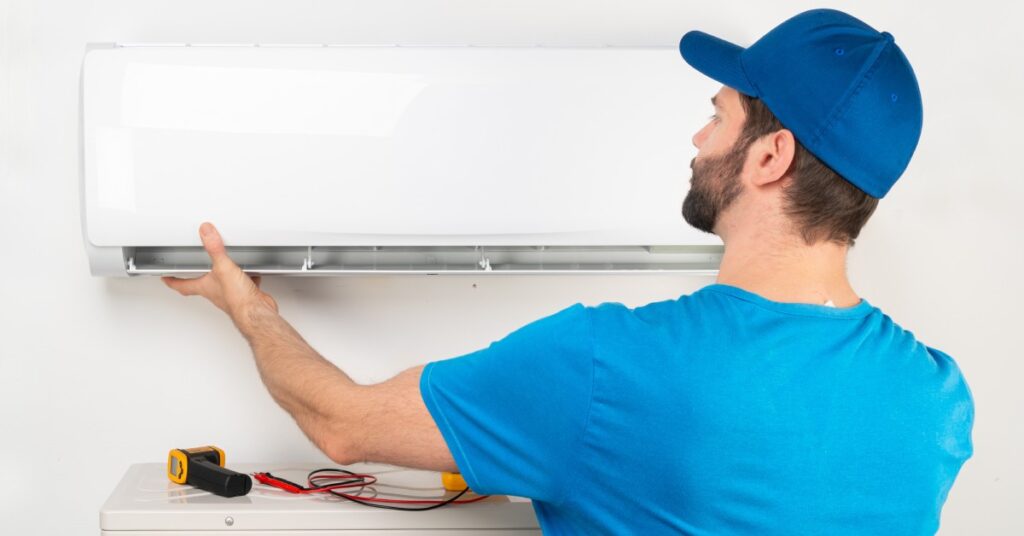 A technician wearing a blue shirt and hat replaces a component on an in-home wall air conditioning unit.