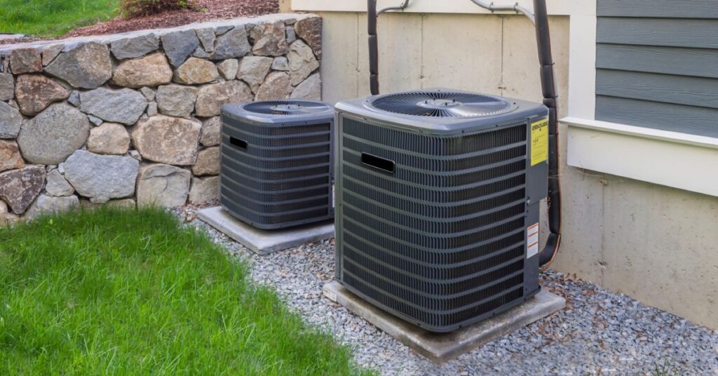 Two HVAC systems installed outside of a home with grey shiplap siding. Greenery and stone surround the systems.