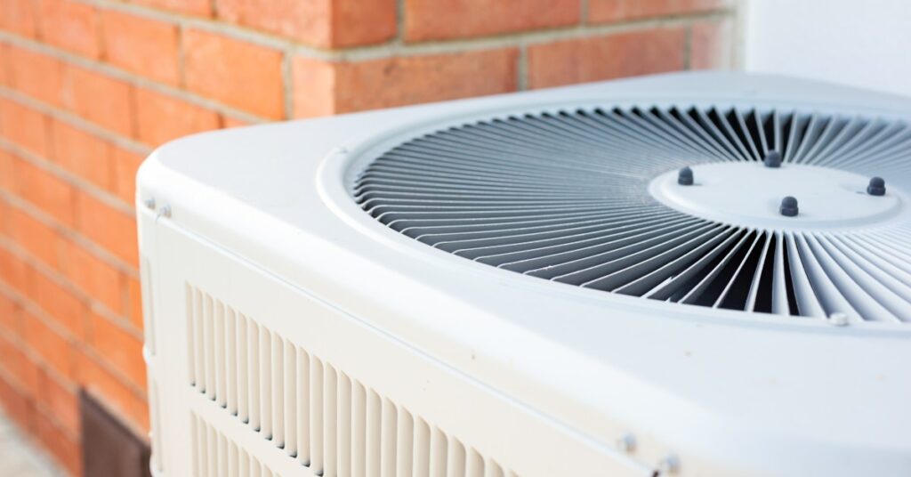 A white air conditioner condenser unit, up close, located outside of a home with brick exterior walls.