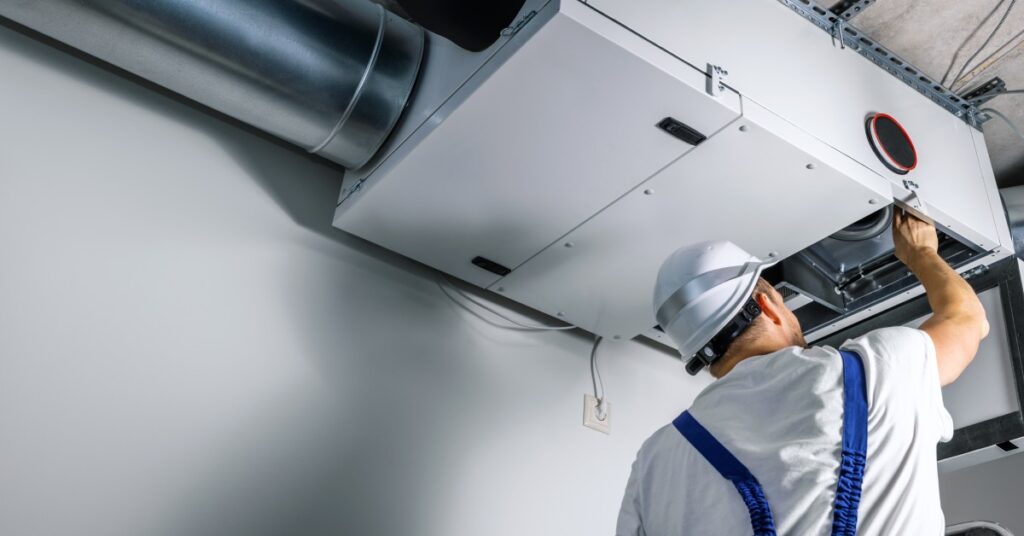 An HVAC engineer wearing a white hard hat and installing a heat recovery ventilation system located on the ceiling of a home.