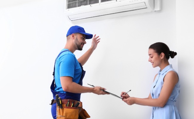 An HVAC service worker wearing blue overalls and a hat holds a clipboard for a woman as she signs the paper on it.