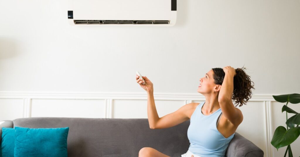 A woman holding up her wavy brown hair looks annoyed as she holds a remote up to her broken air conditioner.