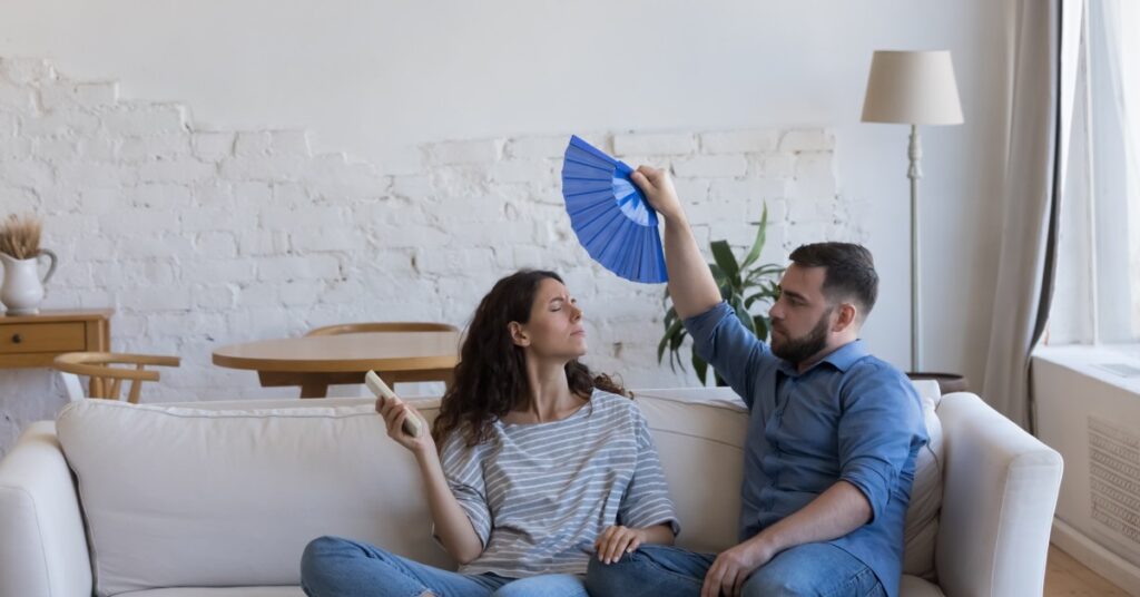 A male and female couple sitting on their couch uncomfortably, fanning each other with a blue folding fan.
