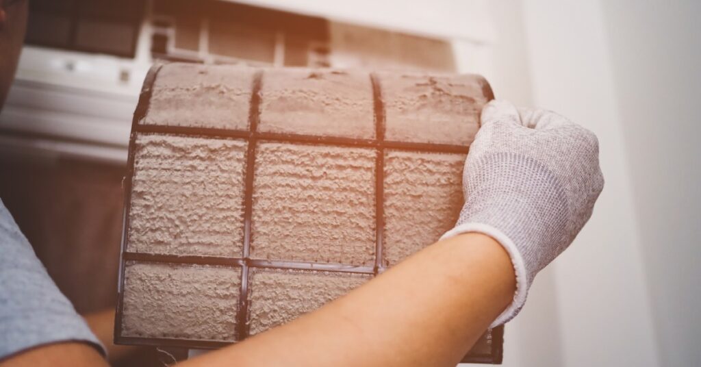 A man wearing gray gloves removes a dusty and clogged air filter from an in-home wall air conditioning unit.