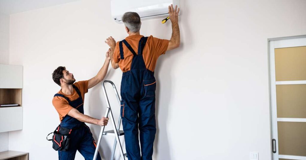 Two repairmen wearing orange shirts and blue overalls inspecting an in-home wall AC unit. One stands on a ladder.