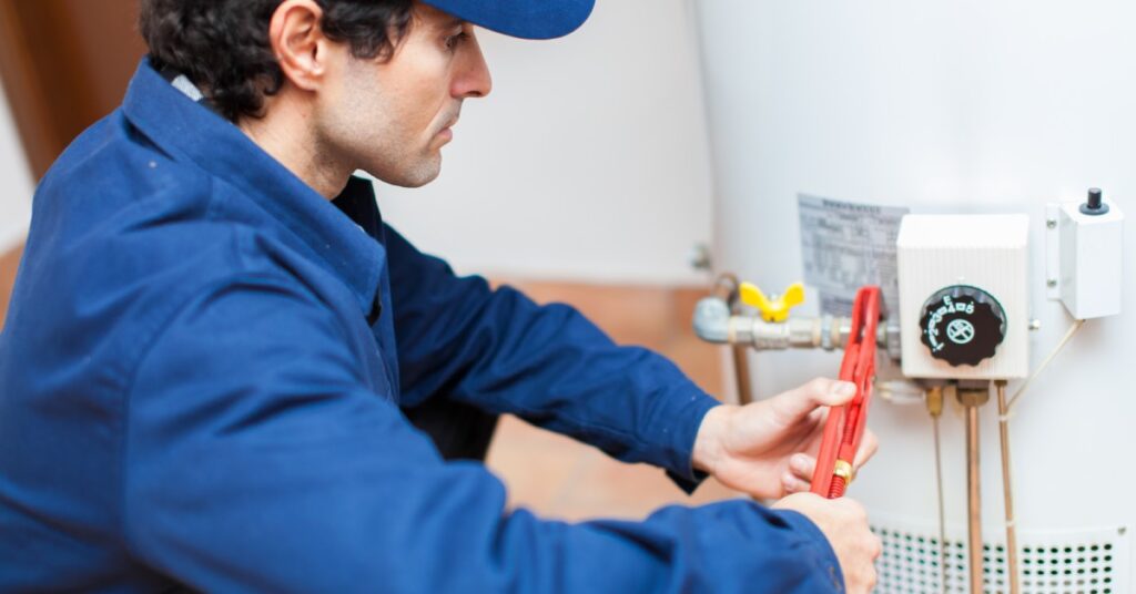 A male service technician crouches down to adjust the pipes around a hot water heater with a wrench.