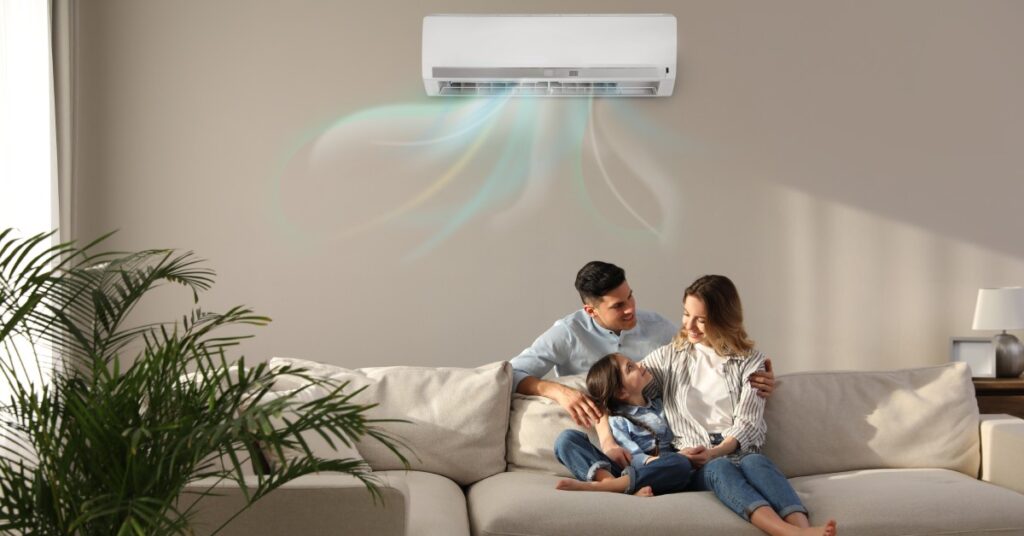 A family sits in the living room of their home on a beige sofa. An air conditioner is installed on the wall behind them.