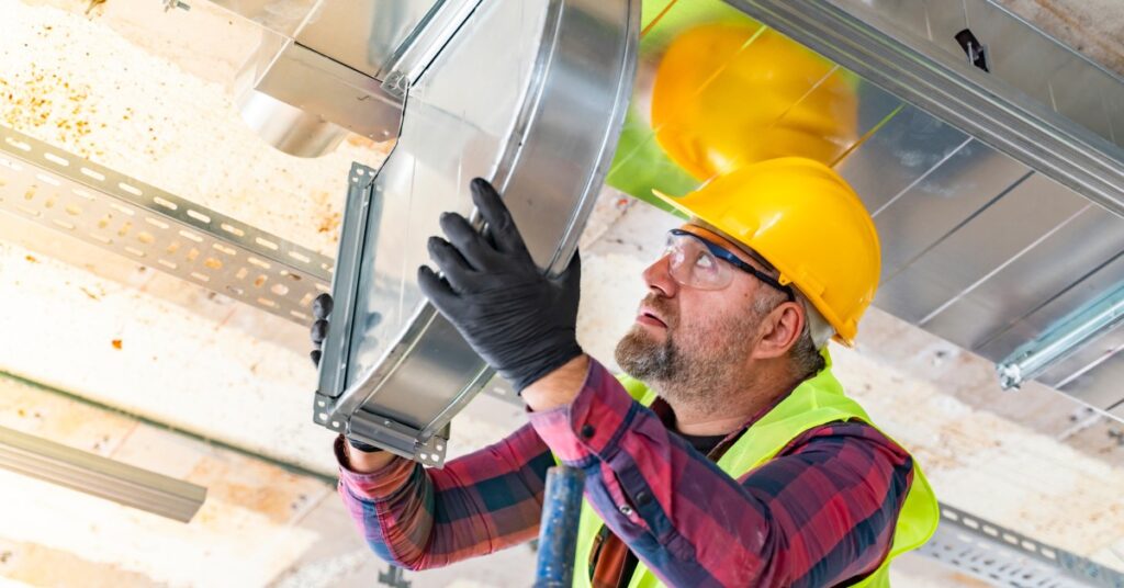 A professional AC service man installs an air conditioning duct while wearing a hard hat and protective eyewear.