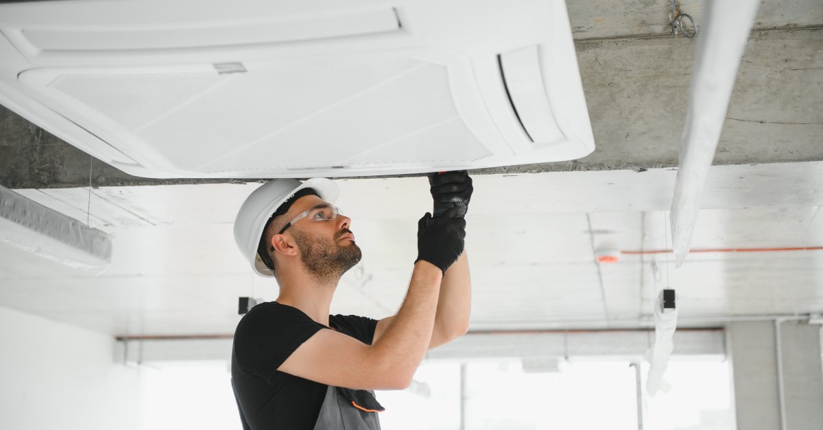 A contractor wearing a hardhat and overalls adjusts a component of an HVAC system on the ceiling of a building.
