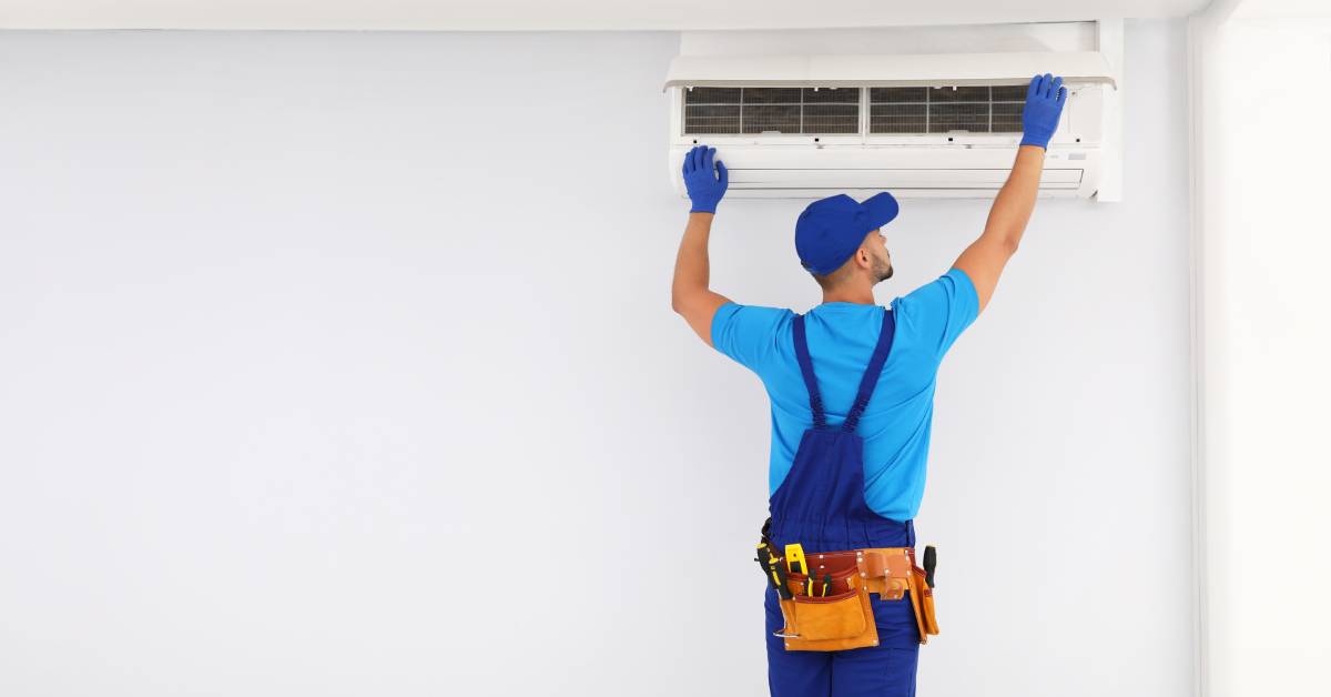 An air conditioning technician in blue overalls and a blue hat standing tall to reach an in-wall AC unit indoors.
