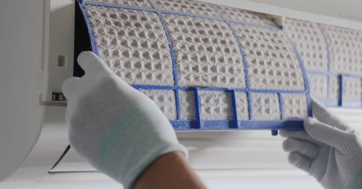 A close-up as two gloved hands remove an air filter clogged with dust from an indoor air conditioning unit.