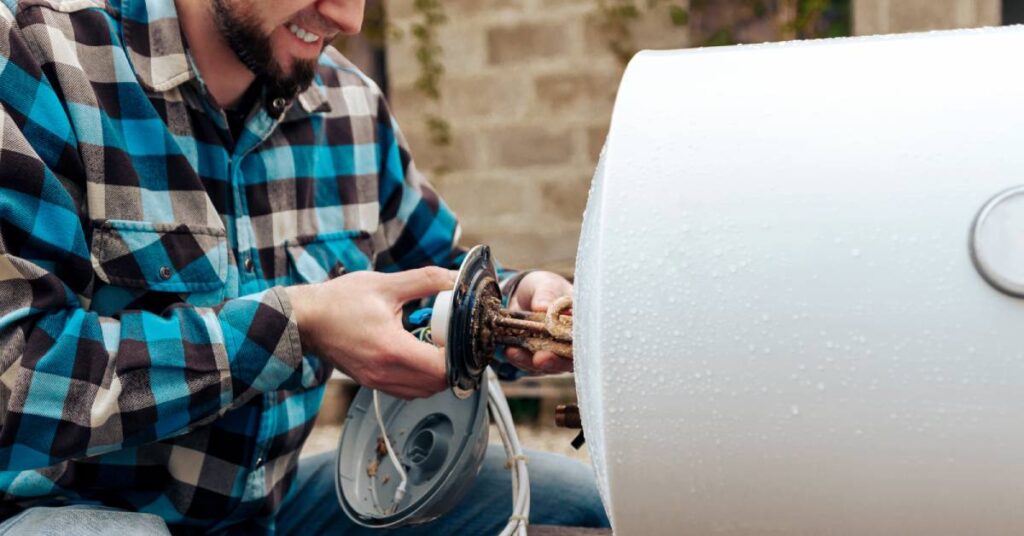 A smiling man wearing a blue plaid shirt removes a component from a hot water heater unit on its side on the ground.