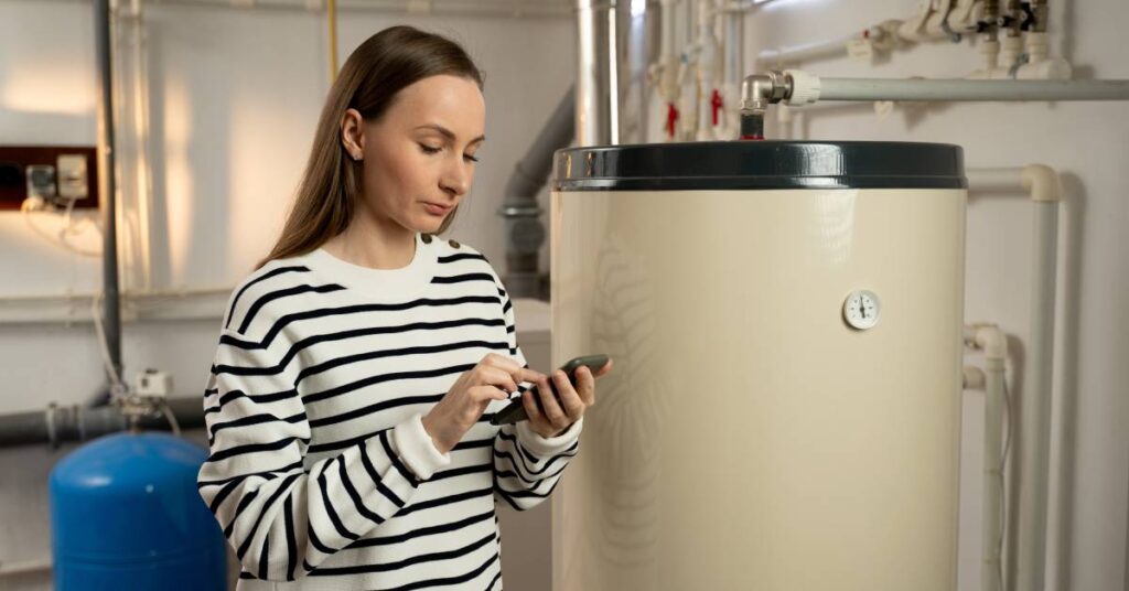 A woman in a striped shirt standing in front of a hot water heater holding her smartphone and tapping the screen.