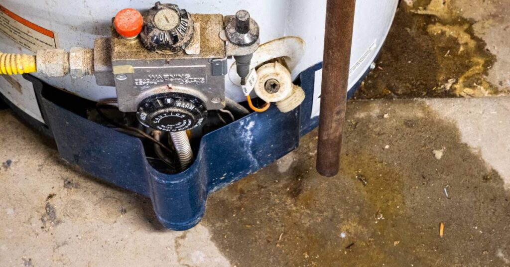 A hot water heater that is leaking onto a concrete floor. The components look older and covered in dust.