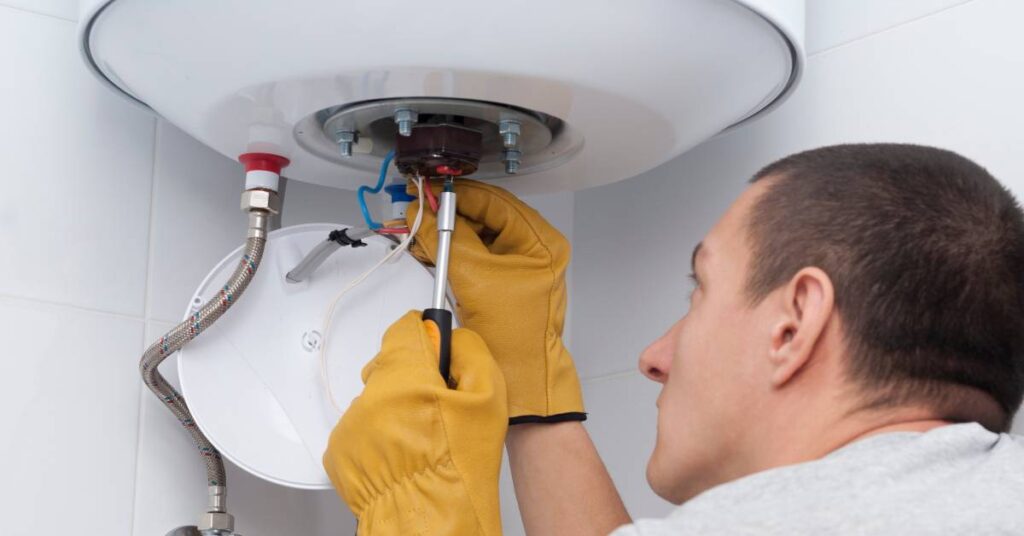 A man with short hair wears yellow gloves reaches and reaches up into the bottom components of a hot water heater.
