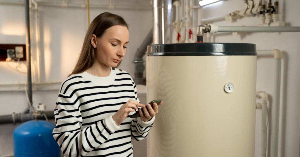A woman in a striped shirt holds a cell phone as she stands in a utility room in front of a hot water heater.