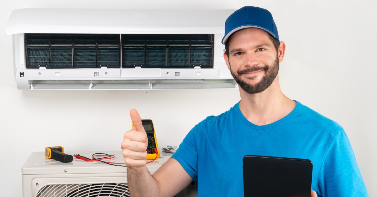 An air conditioner repairman wearing a blue hat and shirt stands in front of a unit and gives a thumbs up.