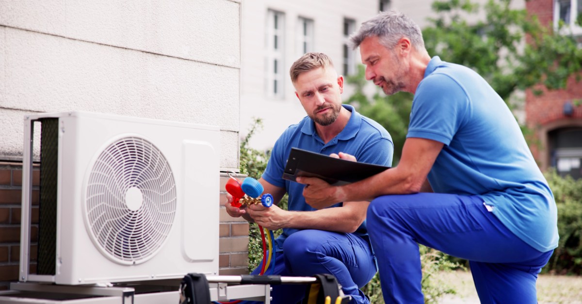 Two AC repairmen kneel down in front of an outdoor unit, working with the wires and discussing repairs.