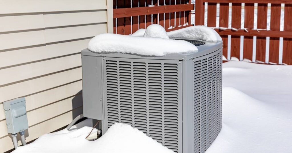A gray outdoor HVAC unit sits covered in a light layer of snow, with a deep layer of snow surrounding it.