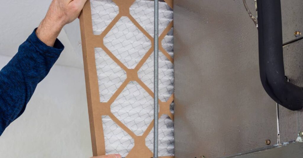 A pair of hands from the left install a clean air filter into an HVAC duct in a building's ceiling.