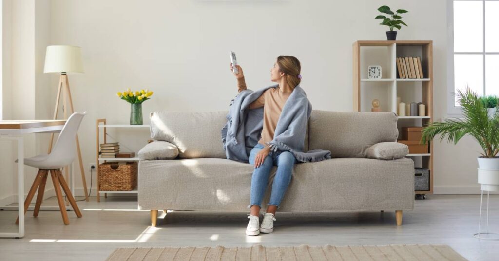 A woman wrapped in a blue blanket sits on her couch and uses a remote to adjust the air conditioner behind her.