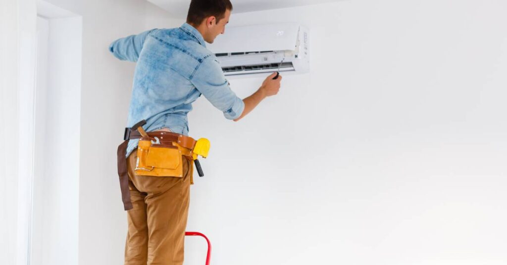 A maintenance worker wearing a toolbelt stands on a ladder to reach an in-unit air conditioner, using a tool to open it.